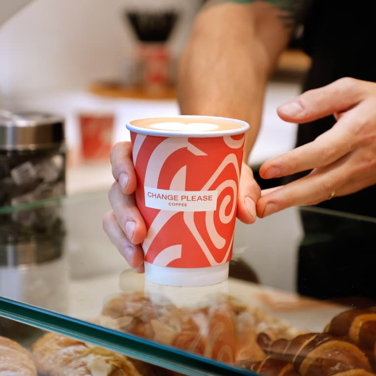 A hand holding a vibrant red coffee cup labeled "Change Please Coffee" over a glass counter displaying pastries.