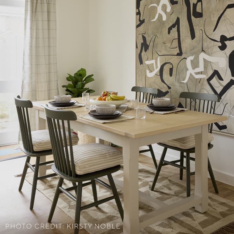 Modern dining room with a light wooden table set for four, featuring striped cushions, abstract wall art, a plant, and a bowl of fruit.