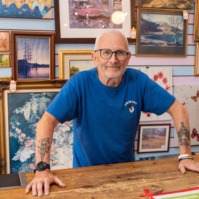 A man in a blue shirt with tattoos stands behind a wooden counter, surrounded by various framed artworks on display.