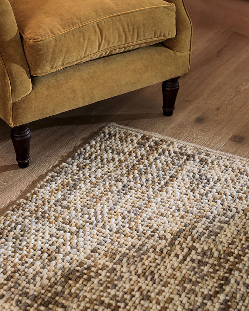 A mustard yellow armchair with dark wooden legs sits on a wooden floor next to a textured, multicolored woven rug.