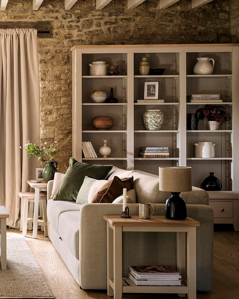 Cosy living room with a beige sofa, green and brown cushions, a wooden side table, bookshelves with decor, and stone walls.