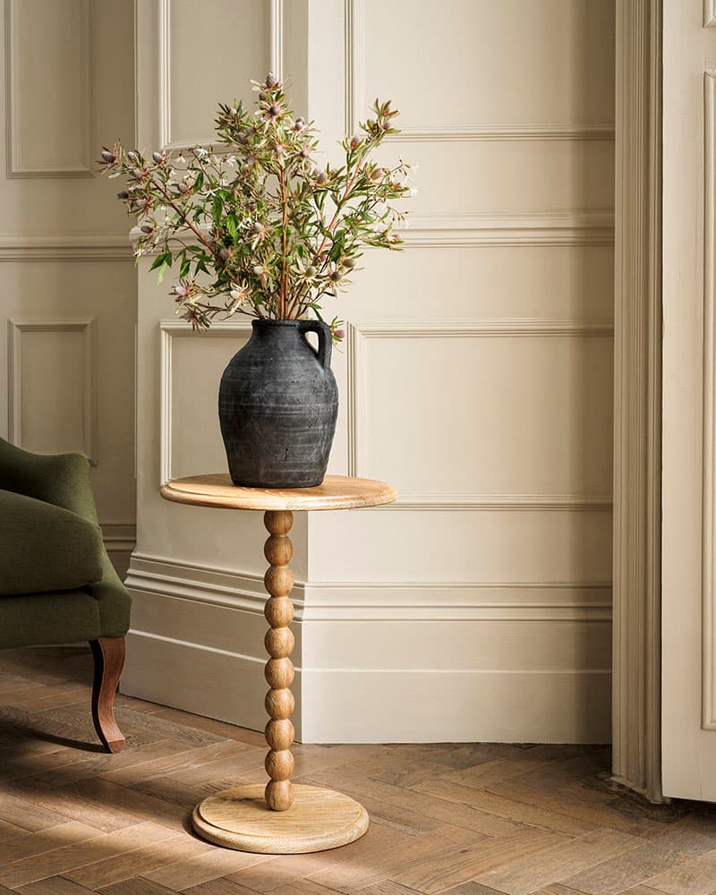 A rustic ceramic jug with branches and leaves on a wooden pedestal table, in a softly lit room with paneled walls and a partial view of a green chair.