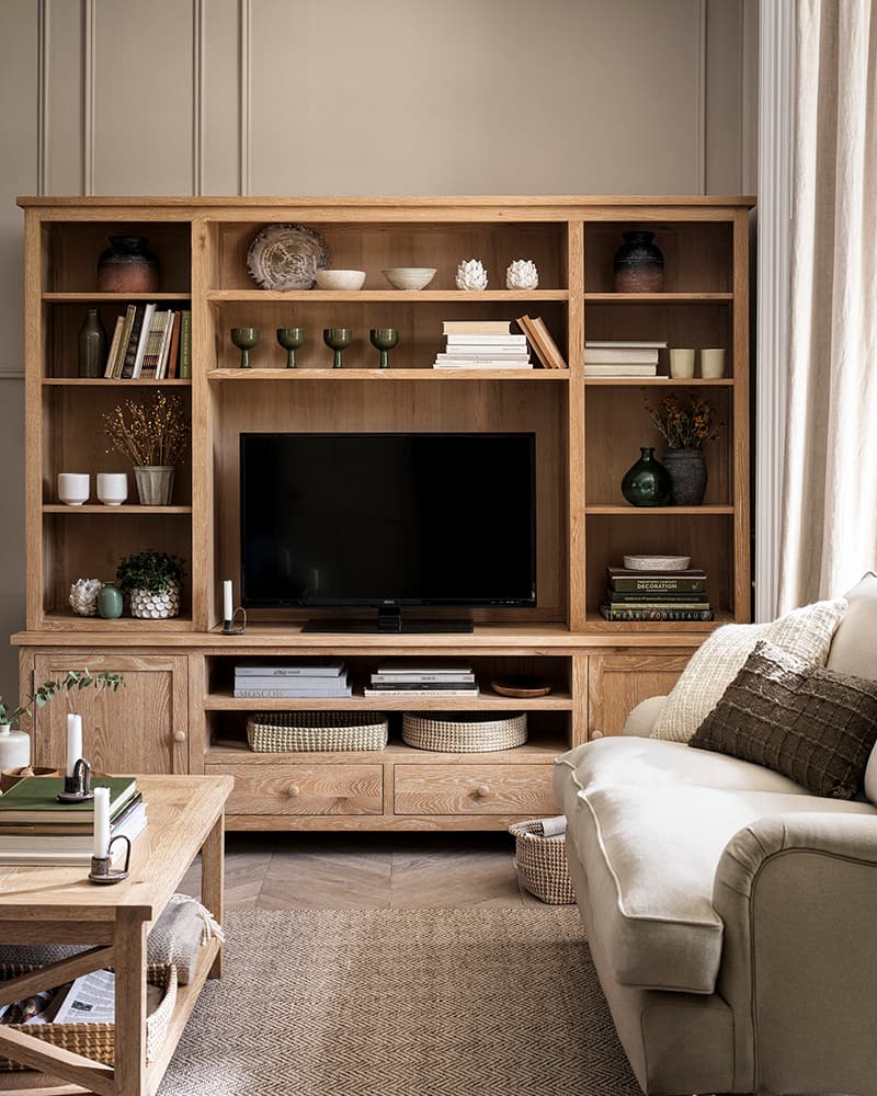 Living room with a wooden entertainment unit, TV, books, decorative items, baskets, and a beige couch with textured cushions.