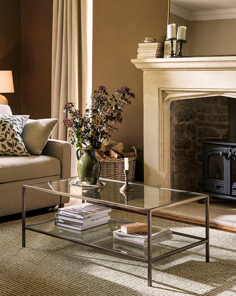 Cosy living room with a beige sofa, glass coffee table, vase of flowers, stack of books, and a stone fireplace with a wood-burning stove.
