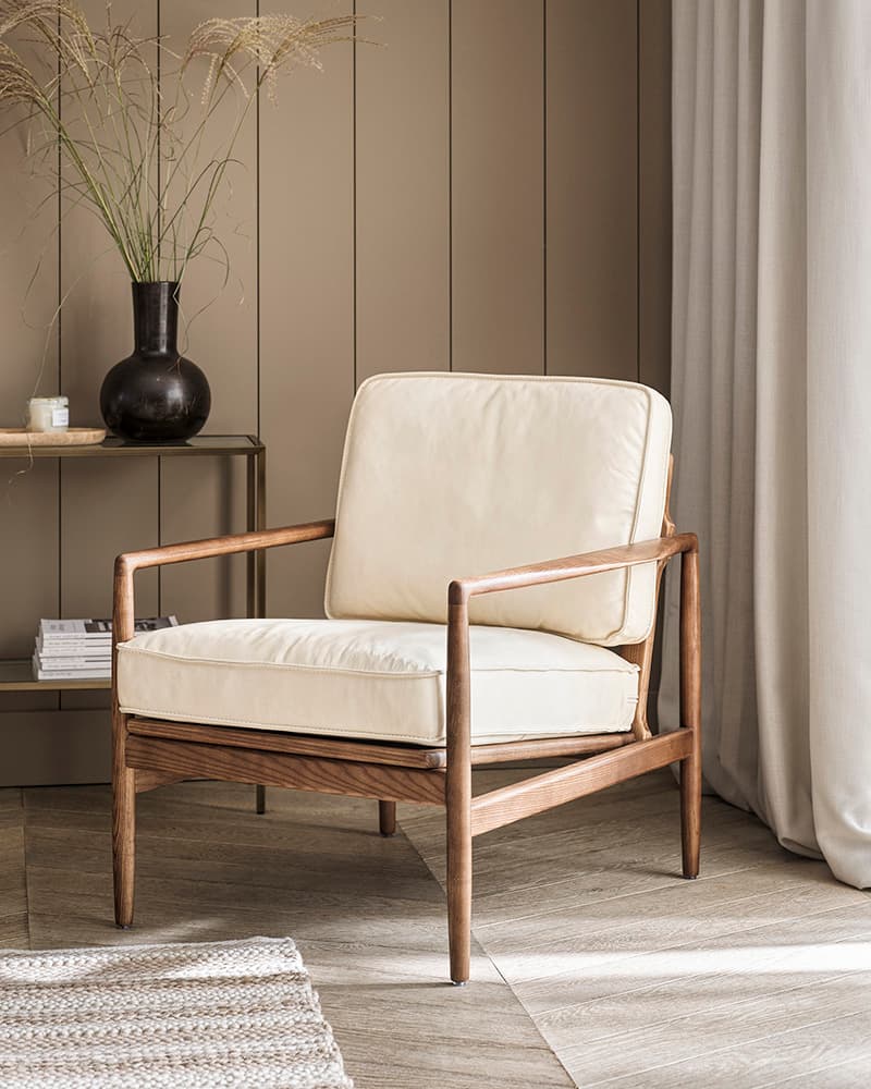 A modern wooden armchair with cream cushions next to a textured rug, a vase of tall grass, and books on a glass side table in a cosy room.