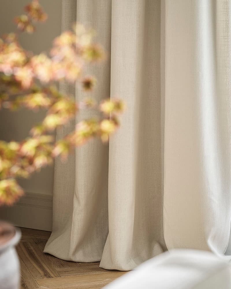 Cream-colored curtains hang in a softly lit room with blurred autumn leaves in the foreground.