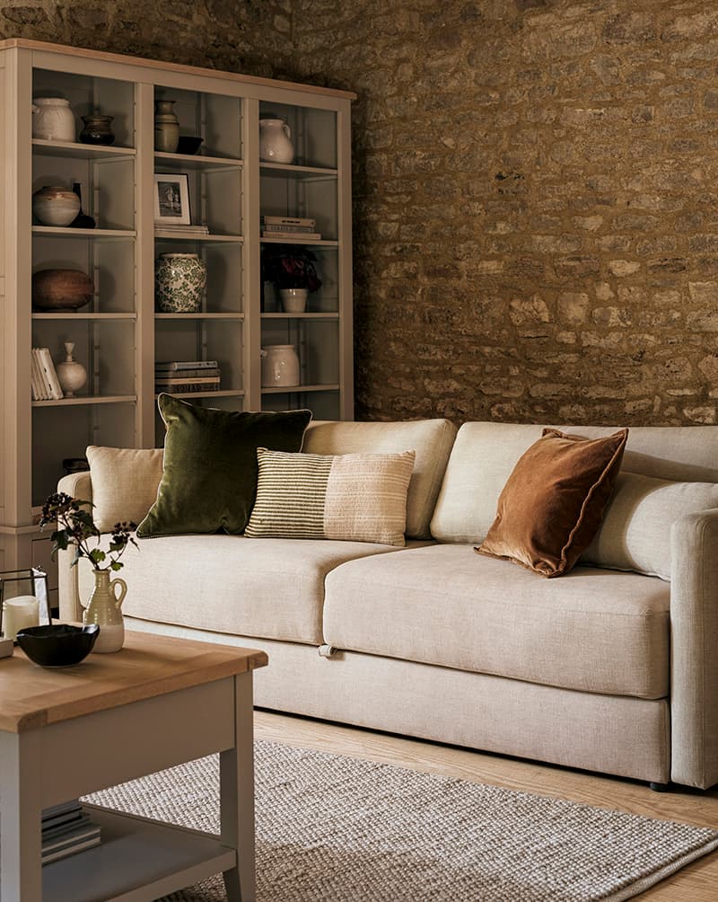 Living room with a beige sofa, colorful cushions, a wooden coffee table, and a shelving unit filled with decor against a stone wall.