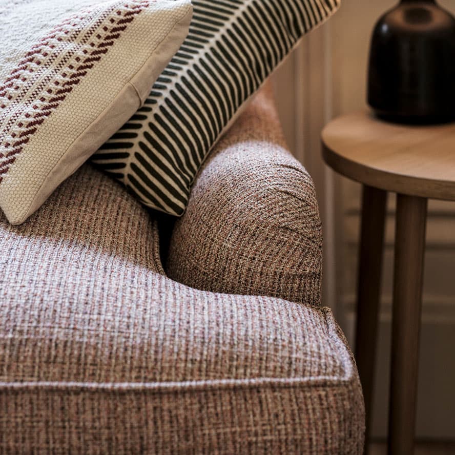 A textured sofa with two patterned cushions beside a round wooden side table and a dark decorative vase.