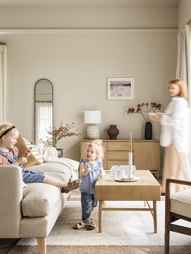 Cozy living room with two children, one on the sofa, the other standing by a coffee table, and a woman walking past, decorated in neutral tones.