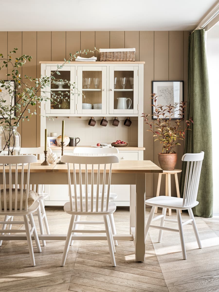 Bright dining room with a wooden table, white chairs, and a hutch. Decor includes green plants, a wicker basket, and wall art.