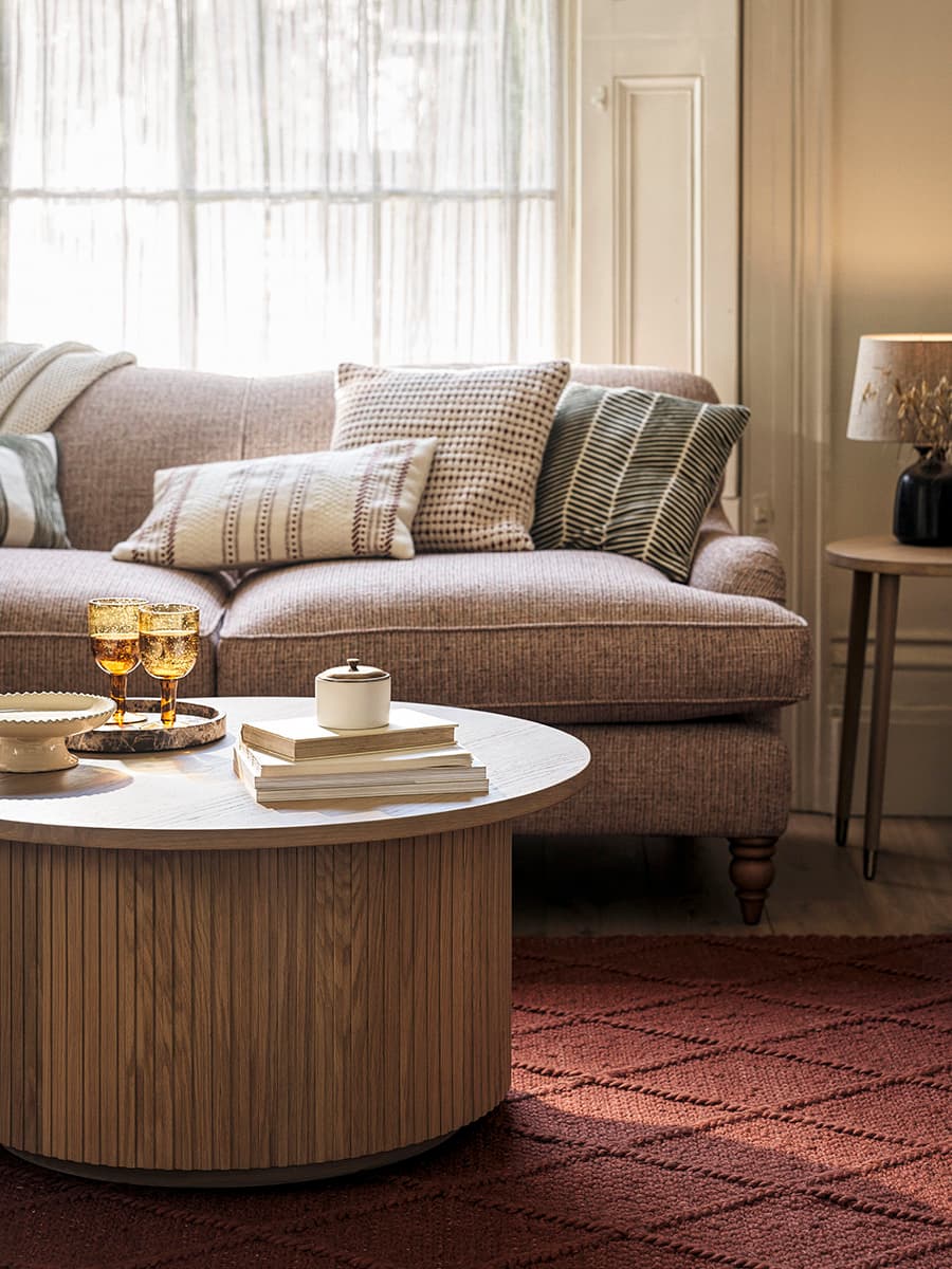 Living room with a textured fabric sofa, assorted cushions, round wooden coffee table with books and glasses, and a red patterned rug.