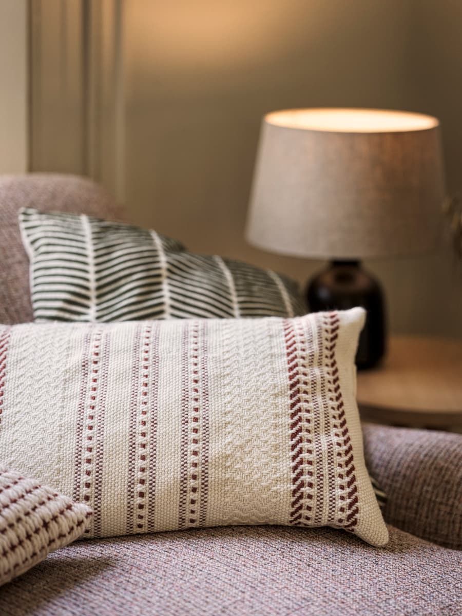 A cosy living room with a textured white pillow with red patterns on a sofa, alongside another pillow with green stripes and a lit table lamp in the background.