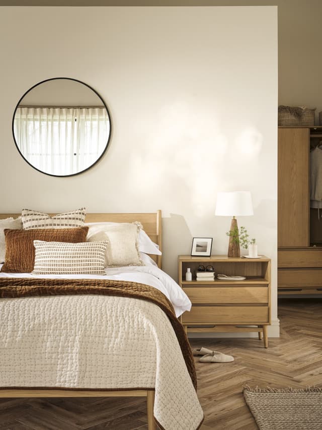 Minimalist bedroom with a wooden bed, neutral bedding, round mirror, wooden nightstand, and soft lighting. Herringbone floor and a cozy atmosphere.