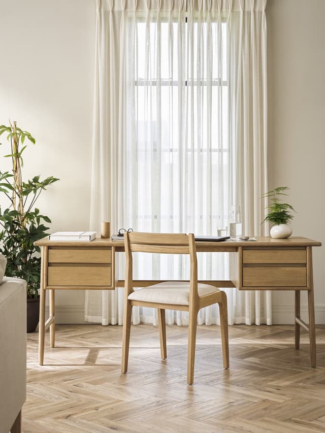 Minimalist home office with a wooden desk and chair, potted plant, and sheer curtains in a bright room with herringbone wood flooring.