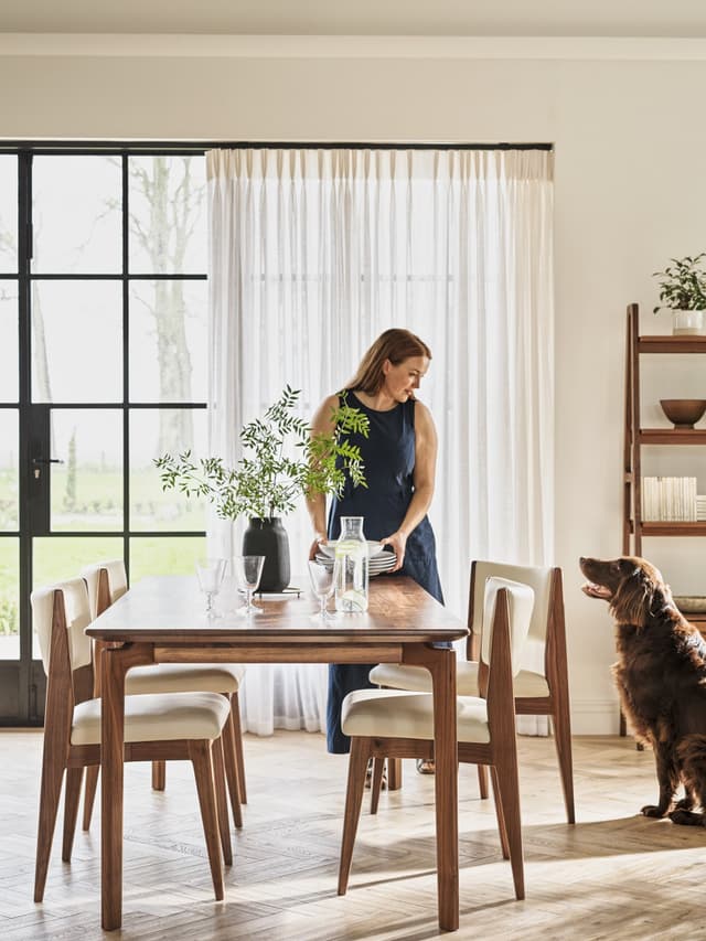 Woman setting a wooden dining table with plants and glassware, a dog nearby, in a bright room with large windows and white curtains.