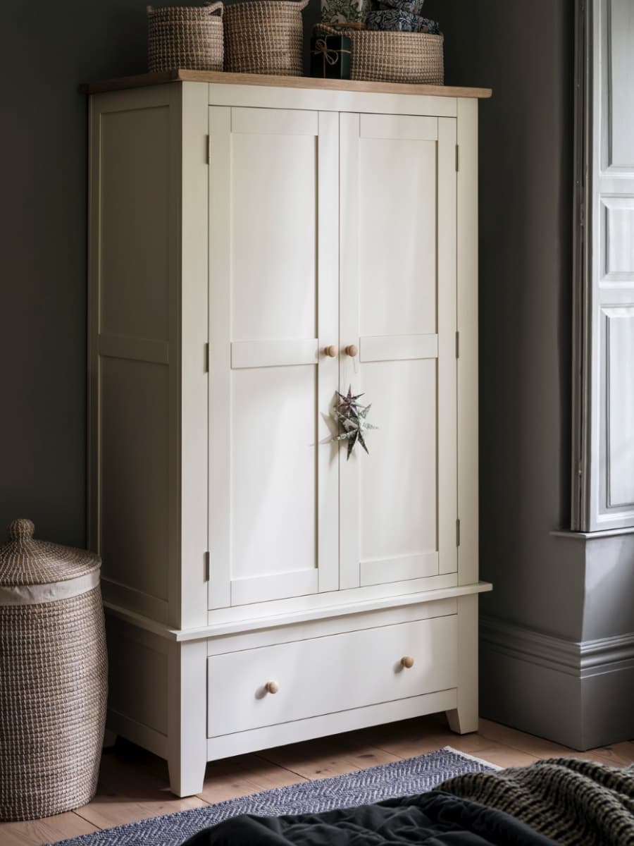 White wooden wardrobe with closed doors and a drawer, adorned with a small hanging plant. Baskets on top, wicker hamper beside, near a window.