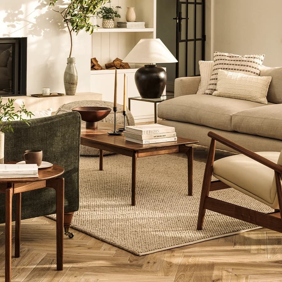A cozy living room with beige sofa, wooden coffee table, potted plants, and a black table lamp. Light streams in creating a warm ambiance.