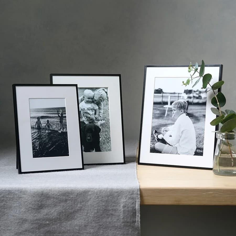 Three black-and-white framed photographs on a wooden surface, one with a plant in a glass jar beside it, set against a grey background.